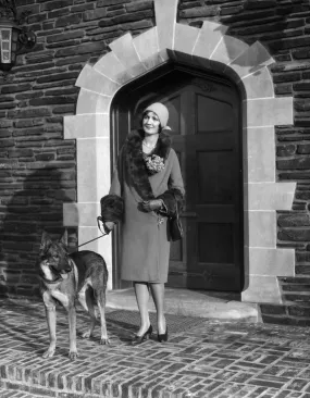1920s fashionable smiling woman wearing cloche hat coat with fur trim and german shepherd dog on leash standing in arched doorway